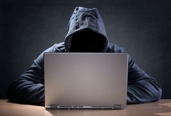 zdjęcie kolorowe: osoba siedząca przed komputerem, ubrana w czarna bluzę dresową z założonym kapturem na głowę