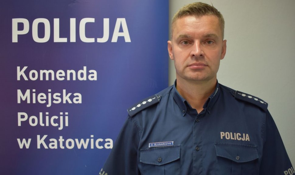 zdjęcie kolorowe: asp. szt. Grzegorz Ślusarczyk na tle granatowego baneru Komendy Miejskiej Policji w Katowicach