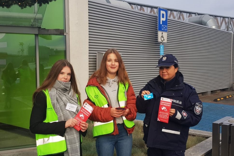 Na kolorowym zdjęciu widać policjantkę z dwoma wolontariuszkami, stoją na parkingu 