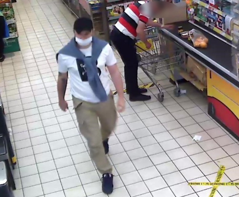 zdjęcie kolorowe: zapis z monitoringu sklepowego przedstawiający mężczyznę podejrzewanego o kradzież alkoholu,  ubrany w beżowe długie spodnie, biała koszulkę z krótkim rękawem i przewieszonym przez ramiona szarym swetrem zawiązanym za rękawki na supeł opadający na klatkę piersiową mężczyzny, czarną czapeczkę z daszkiem i białą maseczkę ochronną założoną na usta. 