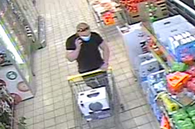zdjęcie kolorowe: zapis z monitoringu sklepowego przedstawiający mężczyznę podejrzewanego o kradzież alkoholu ubrany w niebieskie długie jeansy, czarna koszulkę typu polo z krótkim rękawem, niebieską maseczkę ochronną założoną na usta i okulary przeciwsłoneczne czarne z błyszczącymi pomarańczowymi szybkami, które założone są na głowie pchający koszyk sklepowy