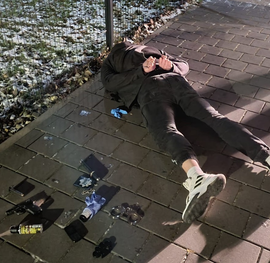 zdjęcie kolorowe: zatrzymany mężczyzna podszywający się za policjanta, leżący na chodniku z rekami skuty kajdankami, obok niego na chodniku leży broń, kajdanki, fałszywa legitymacja, gaz