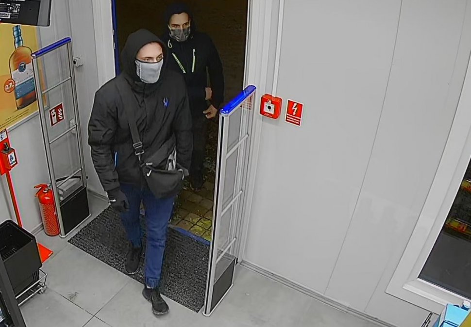 zdjęcie kolorowe: dwóch mężczyzn ubranych w ciemne ubrania z maseczkami ochronnymi na twarzy wchodzącymi do sklepu