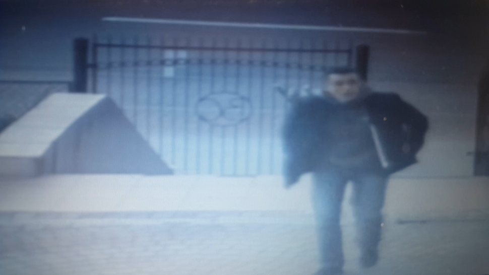zdjęcie kolorowe: zdjęcie z zapisu monitoringu przedstawiające mężczyznę ubranego w ciemne ubrania na tle bramy idący chodnikiem