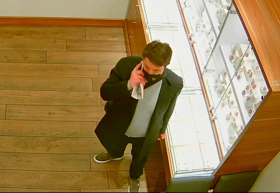 zdjęcie kolorowe: screen z monitoringu sklepowego przedstawiający mężczyznę ubranego w ciemne spodnie, ciemny płaszcz, szary sweter z założona czarna maseczka ochronna na twarzy i rozmawiający przez telefon komórkowy, widok z przodu
