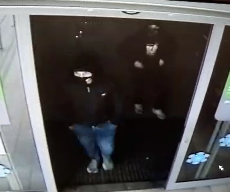 zdjęcie kolorowe: dwóch mężczyzn stojących w drzwiach marketu, pierwszy ubrany w niebieskie spodnie, czarną kurtkę i czarna czapkę oraz założoną na twarzy maseczka ochronną, drugi z mężczyzn ubrany w czarne spodnie, czarna kurtkę i z maseczka ochronną na twarzy