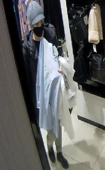 zdjęcie kolorowe: zrzut obrazu z monitoringu sklepowego przedstawiający kobietę w czarnej kurtce, ciemnych spodniach i szarych butach, szarej wełnianej czapce gdzie na twarzy ma założoną maseczkę ochronną a w ręce trzyma ubrania na wieszakach