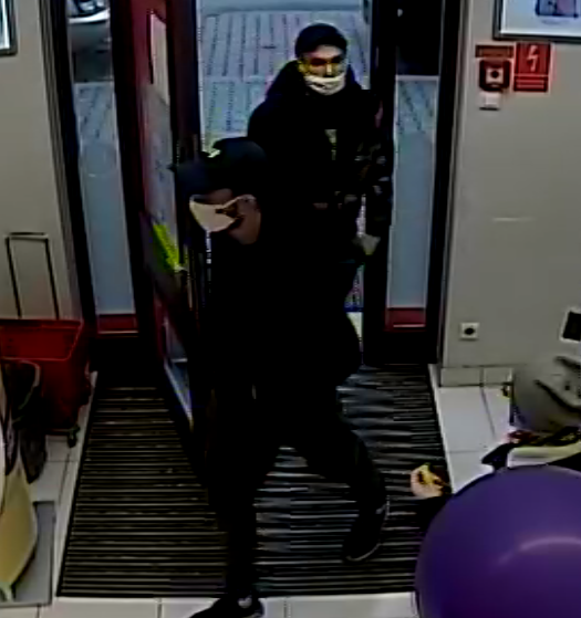 zdjęcie kolorowe: dwóch mężczyzn ubranych w czarne ubrania z czapeczkami na głowie i maseczkami ochronnymi na twarzy wchodzący do sklepu