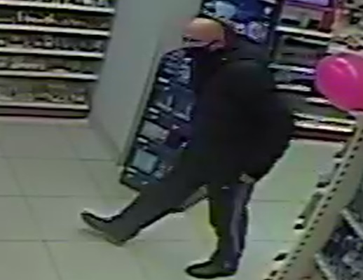 zdjęcie kolorowe: zrzut ze sklepowego monitoringu przedstawiający przygarbionego łysego mężczyznę ubranego w czarne buty, czarne spodnie, czarna kurtkę i czarna maseczkę ochronna na twarzy w sklepie kosmetycznym