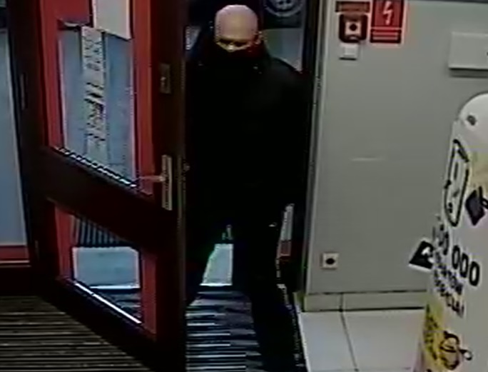 zdjęcie kolorowe: zrzut ze sklepowego monitoringu przedstawiający łysego mężczyznę ubranego w czarne buty, czarne spodnie, czarna kurtkę i czarna maseczkę ochronna na twarzy wchodzącego do sklepu