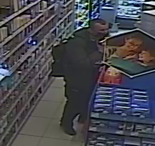 zdjęcie kolorowe: zrzut z monitoringu sklepowego przedstawiający mężczyznę w ciemnozielonej kurtce, ciemnych spodniach i czarnych butach z maseczka ochronną na twarzy oglądający towar na półkach