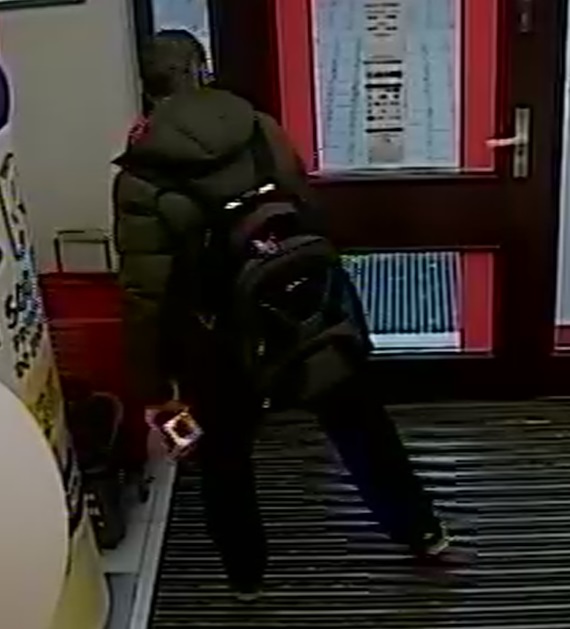 zdjęcie kolorowe: zrzut z monitoringu sklepowego przedstawiający mężczyznę w ciemnozielonej kurtce, ciemnych spodniach i czarnych butach z maseczka ochronną na twarzy i perfumami trzymanymi w dłoni, kiedy wychodzi ze sklepu