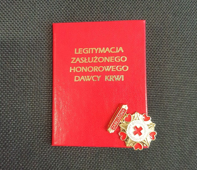 zdjęcie kolorowe: legitymacja odznaczenia „Zasłużony Honorowy Dawca Krwi I Stopnia” oraz złota odznaka przedstawiająca czerwony krzyż obtoczony czerwonymi serduszkami dla Pana Dawida Korycińskiego