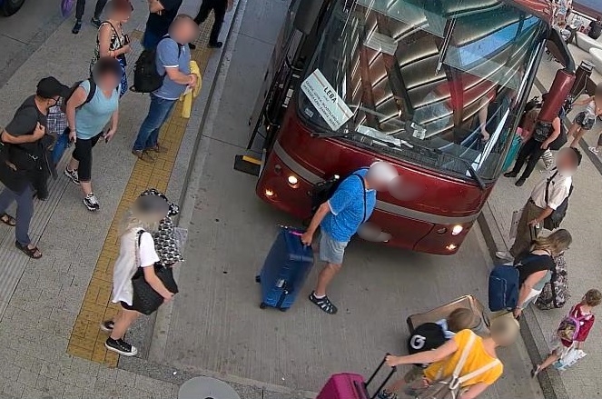 zdjęcie kolorowe: dworzec autobusowy, grupa ludzi oczekująca na peronie przed autobusem i podejrzewany o kradzież kieszonkową mężczyzna ubrany był w ciemnoszarą koszulkę z krótkim rękawem, ciemne spodnie, sandały i czapkę z daszkiem koloru czarnego, który na prawym ramieniu trzyma torbę