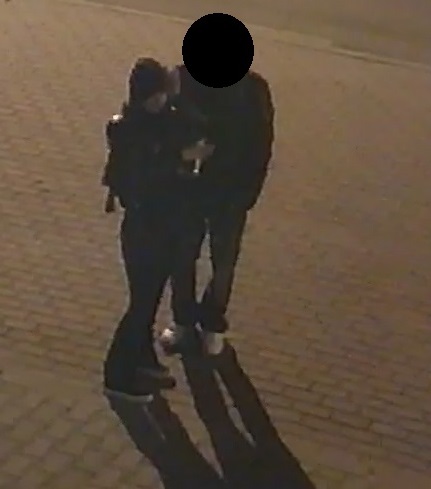 zdjęcie czarno białe: zrzut z monitoringu przedstawiający osobę idącą chodnikiem, ubraną w ciemne spodnie, ciemną bluzę z kapturem założoną na głowę i czarna kurtkę w towarzystwie drugiej osoby