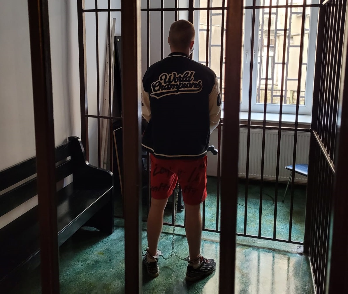 zdjęcie kolorowy: podejrzany zatrzymany za rozbój z założonymi kajdankami zespolonymi czekający w pomieszczeniu dla osób zatrzymanych