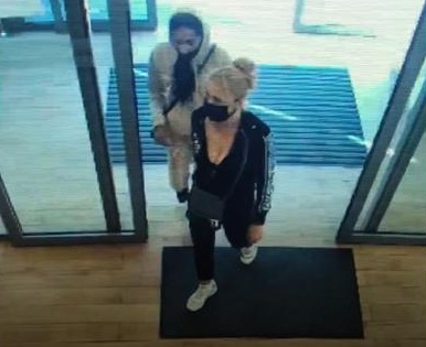 zdjęcie kolorowe: dwie osoby wchodzące przez hotelowe drzwi do budynku: kobieta w blond włosach i w czarnej bluzę i czarnych spodniach z maseczką ochronna na twarzy oraz osoba ubrana w beżowe spodnie, beżową kurtkę, czarną bluzę