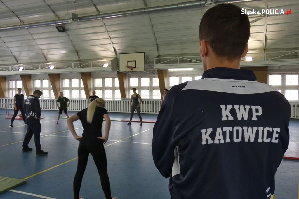 zdjęcie kolorowe: policjant w granatowym dresie z napisem na plecach o treści KWP Katowice, obserwujący osoby ćwiczące na hali sportowej
