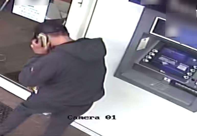 zdjęcie kolorowe: zdjęcie kolorowe: mężczyzna ubrany w czarne spodnie, granatowa kurtkę z kapturem i granatową czapkę z daszkiem, rozmawia przez telefon komórkowy stojąc przy bankomacie