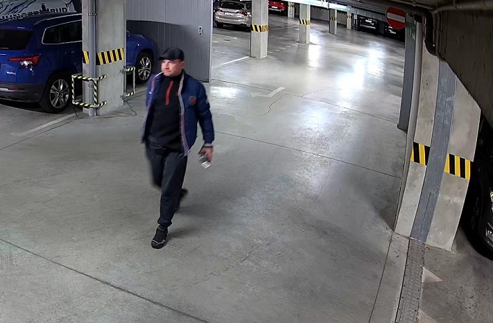 zdjęcie kolorowe: mężczyzna idący przez parking podziemny. Mężczyzna ubrany jest w ciemne buty sportowe, ciemne spodnie, ciemna bluzę sportowa i granatowa kurtkę puchową i ciemno szarą czapkę z daszkiem.