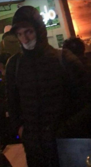 zdjęcie kolorowe: mężczyzna ubrany w czarna kurtkę z kapturem z maseczka założoną na twarzy stojący w lokalu gastronomicznym 