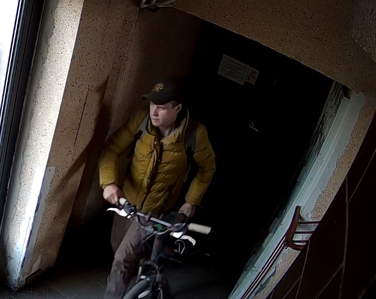 zdjęcie kolorowe: młody mężczyzna ubrany w piaskowa kurtkę puchowa i czapeczkę z daszkiem prowadzący rower w korytarzu