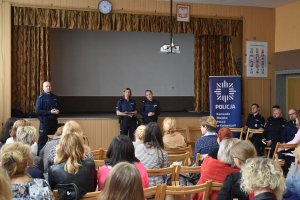 Na zdjęciu widać naczelnika wydziału prewencji KMP Katowice jak mówi do zebranych na auli pedagogów którzy siedzą na krzesłach, obok naczelnika stoją dwie policjantki.