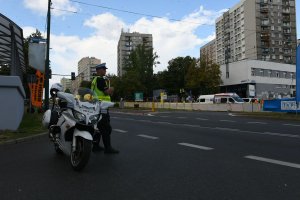 na zdjęciu widać motocykl policyjny obok stoi policjant a za policjantem widoczne są budynki