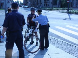 Na zdjęciu widać policjanta i strażnika miejskiego obok stoi rowerzysta w tle widać ulicę