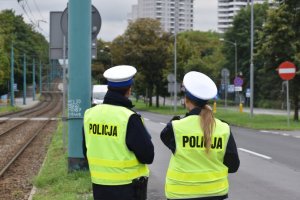 Na zdjęciu widać policjanta i policjantkę ruchu drogowego jak stoją przy drodze. Policjanci mają ubrane kamizelki odblaskowe. W tle widać osiedle Tysiąclecia