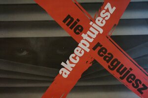 Zdjęcie kolorowe: plakat przedstawiający żaluzje zza których wygląda człowiek, na plakacie na czerwonym tle widnieje  biało-czarny napis: nie reagujesz akceptujesz