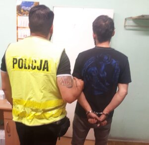 zdjęcie kolorowe: policjant w odblaskowej kamizelce z napisem &amp;quot;Policja&amp;quot; z zatrzymanym mężczyzną