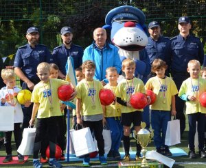 Na zdjęciu widać dzieci oraz nauczycieli, policjantów i maskotkę sznupka jak stoją i pozują do zdjęcia, przed nimi stoją puchary - zdjęcie kolorowe