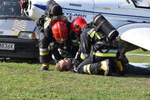 zdjęcie kolorowe: zdjęcie kolorowe: ranna osoba leżąca pod samolotem na płycie lotniska i dwóch strażaków podczas akcji ratowniczej