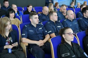 Na kolorowym zdjęciu widać uczestników konferencji siedzących na krzesłach w auli z bok siedzi Pani dziennikarka z TVP Katowice.