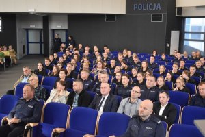 Na kolorowym zdjęciu widać uczestników konferencji siedzących w auli na krzesłach w tel widać napis Policja i drzwi wyjściowe z auli