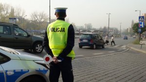 Na zdjęciu widac policjanta ruchu drogowego obok radiowozu który trzyma tarczę do zatrzymywania pojazdów. Przed nim widac ulicę oraz przejście dla pieszych