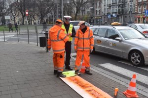 Na zdjęciu widać napis odłóż telefon i żyj naniesiony na przejście dla pieszych obok stoi policjant oraz dwóch pracowników MZUiM