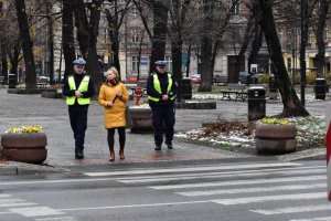 Na kolorowym zdjęciu widać dwóch policjantów ruchu drogowego obok przejścia dla pieszych jak stoją i rozmawiają z przechodniem