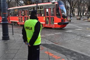 Na kolorowym zdjęciu widać policjanta stojącego w kamizelce odblaskowej w tle widać jadący tramwaj