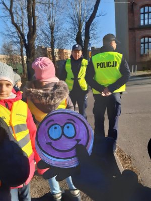 Na kolorowym zdjęciu widać policjanta i dzieci na pierwszym planie w ręce trzymana jest laurka