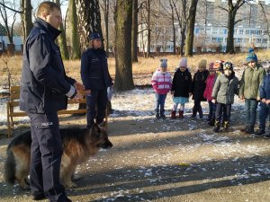 Na zdjęciu widać policjantkę i policjanta którzy stoją wraz z psem na dworze i mówią do grupy przedszkolaków