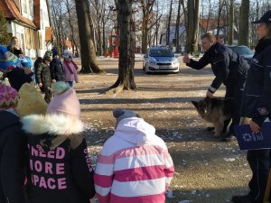 Na zdjęciu widać policjantkę i policjanta którzy stoją na dworze wraz  psem i przedszkolakami