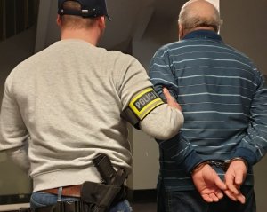 Na zdjęciu widać nieumundurowanego policjanta z opaską policja i pistoletem w kaburze oraz zatrzymanego starszego mężczyznę sprawcę uszkodzenia ciała. Stoją obok siebie