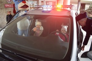 Na zdjęciu widać policjanta z maskotką sznupkiem i dzieci siedzące w radiowozie i stojące obok, radiowóz stoi przed wejściem do przedszkola