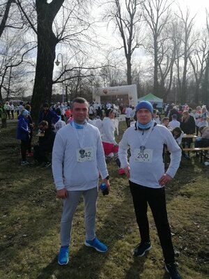 Na zdjęciu widać dwóch policjantów w stroju sportowym w tyle widać uczestników biegu którzy stoją pomiędzy drzewami w parku