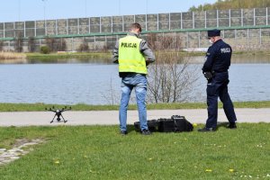 Na zdjęciu widać dwóch policjantów w parku oraz wypuszczonego drona, w tle widać jezioro
