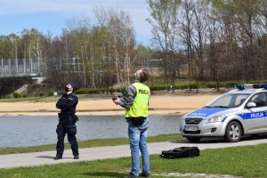 Na zdjęciu widać dwóch policjantów w parku obok stoi radiowóz w tle widać jezioro
