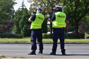 Na zdjęciu widać dwóch policjantów w kamizelkach odblaskowych, którzy stoją przy drodze i mierzą prędkość.