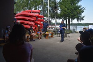 Na zdjęciu widać dzielnicowego i policjantkę którzy stoją na placu i rozmawiają z harcerzami którzy siedzą na ławkach, z tyłu widać czerwone łodzie na stojaku